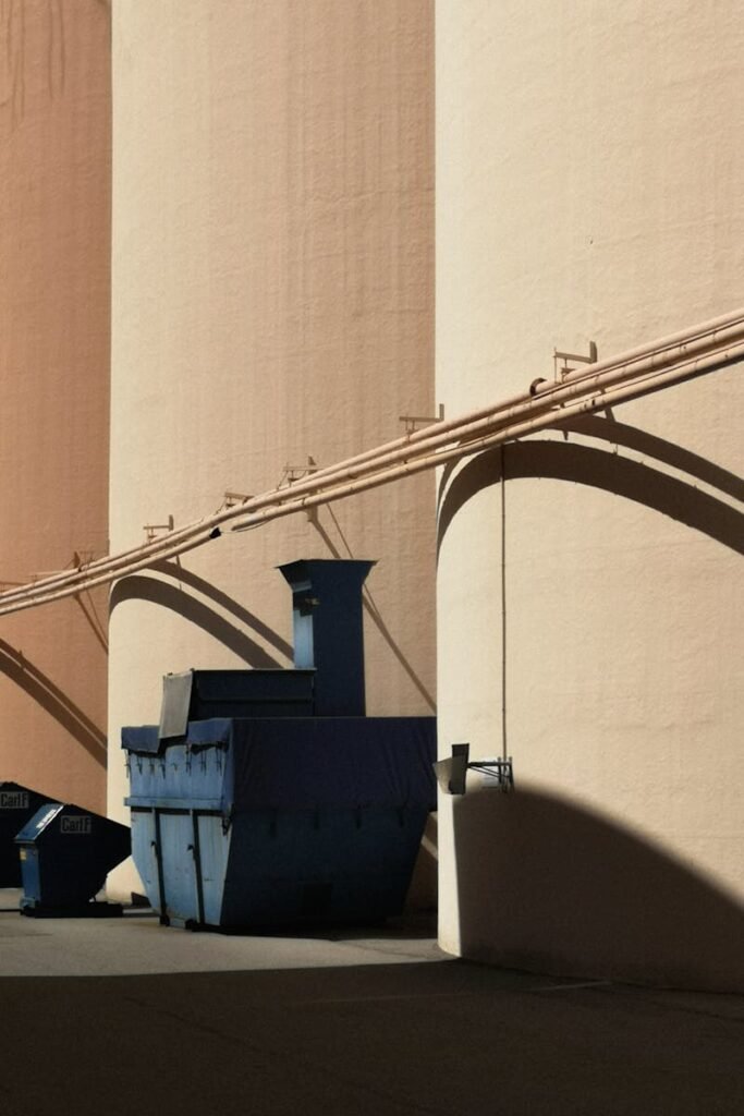 Blue industrial waste containers positioned against a large beige warehouse wall, casting shadows.