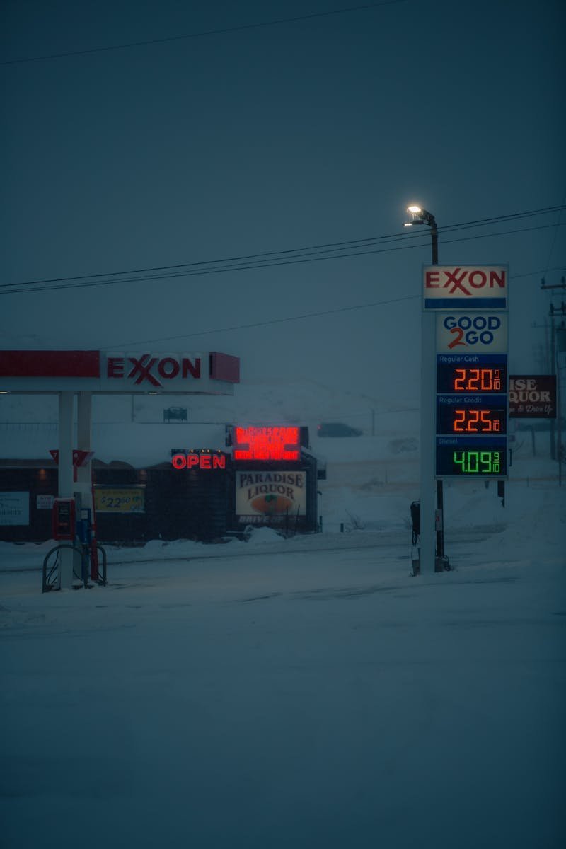 A snowy winter scene of an Exxon gas station with illuminated price signs at dusk.