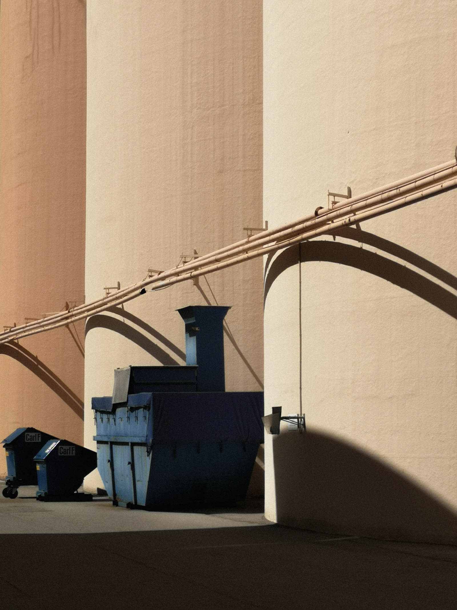 Blue industrial waste containers positioned against a large beige warehouse wall, casting shadows.