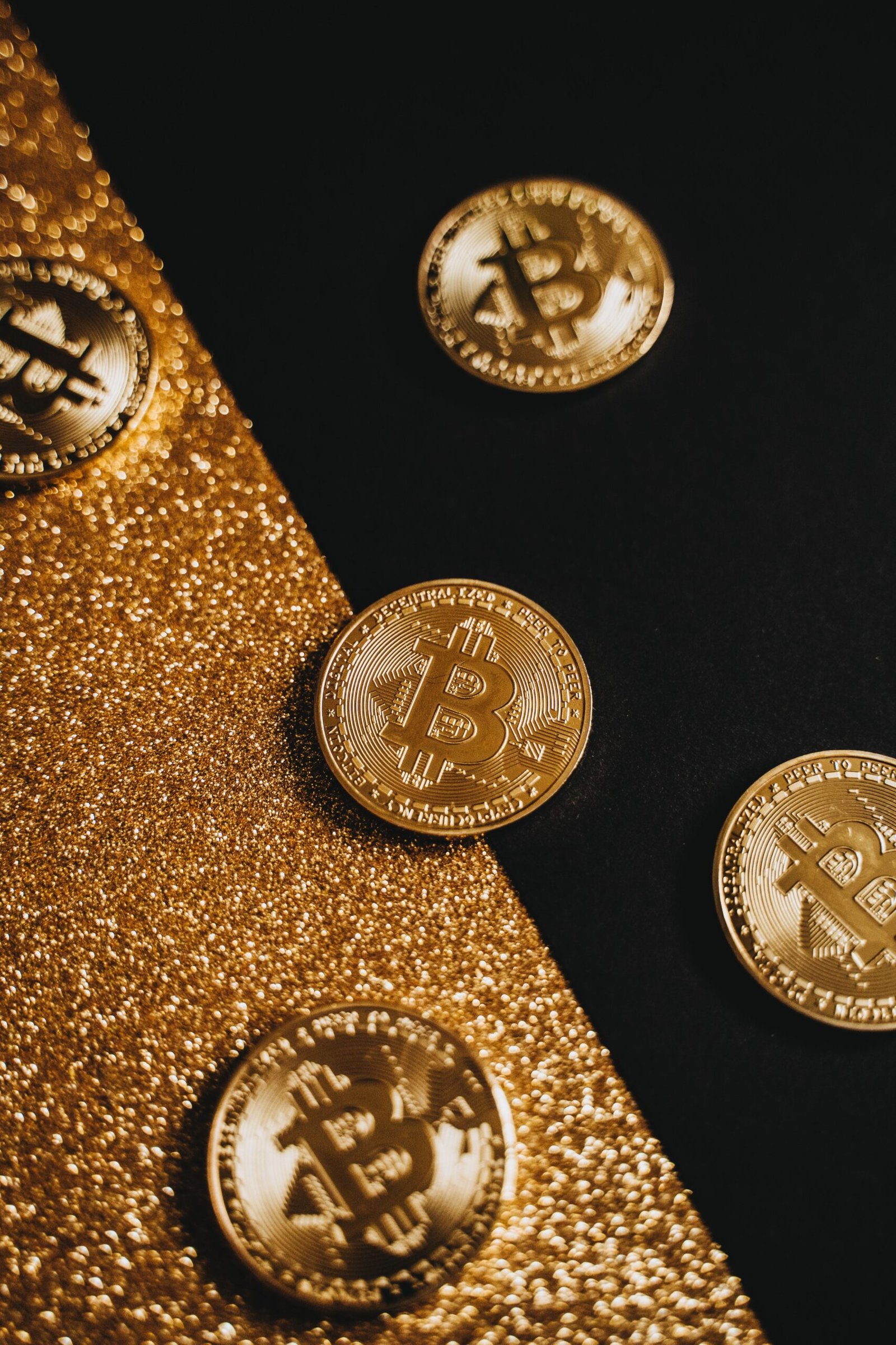 Shiny gold bitcoin coins lying on a split golden glitter and black surface.