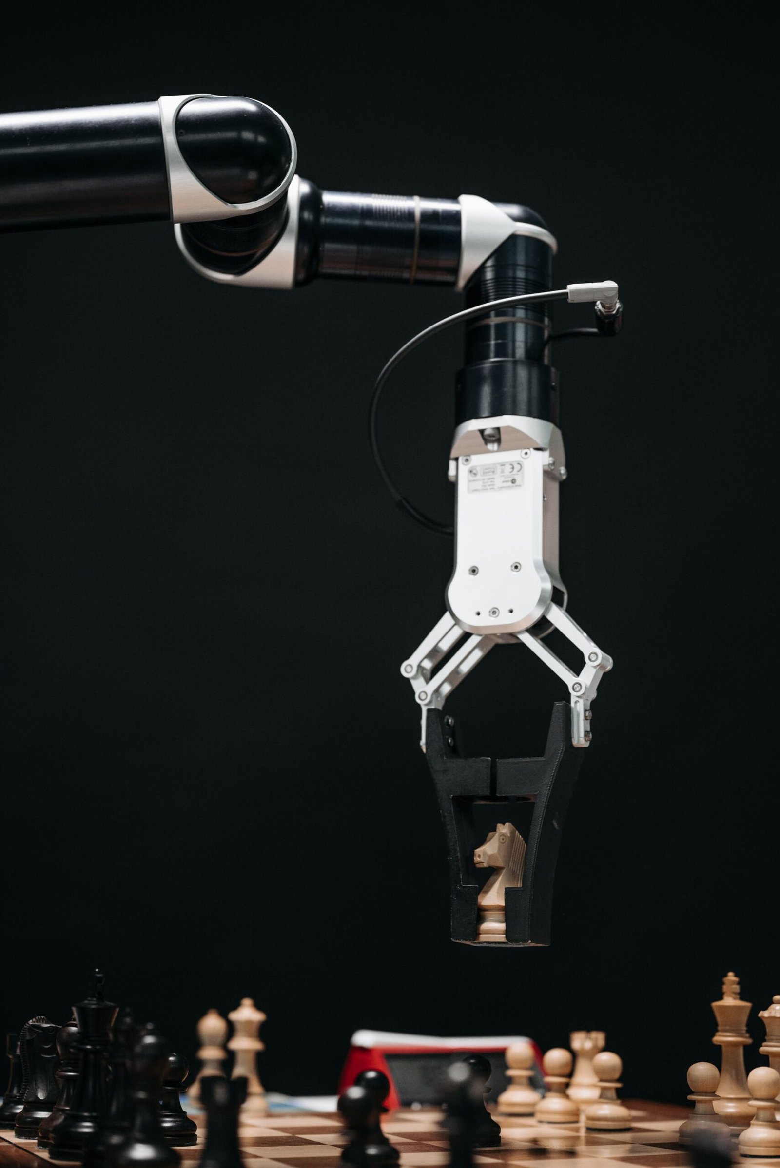 A robotic arm with a pincers holding a knight chess piece on a chessboard.
